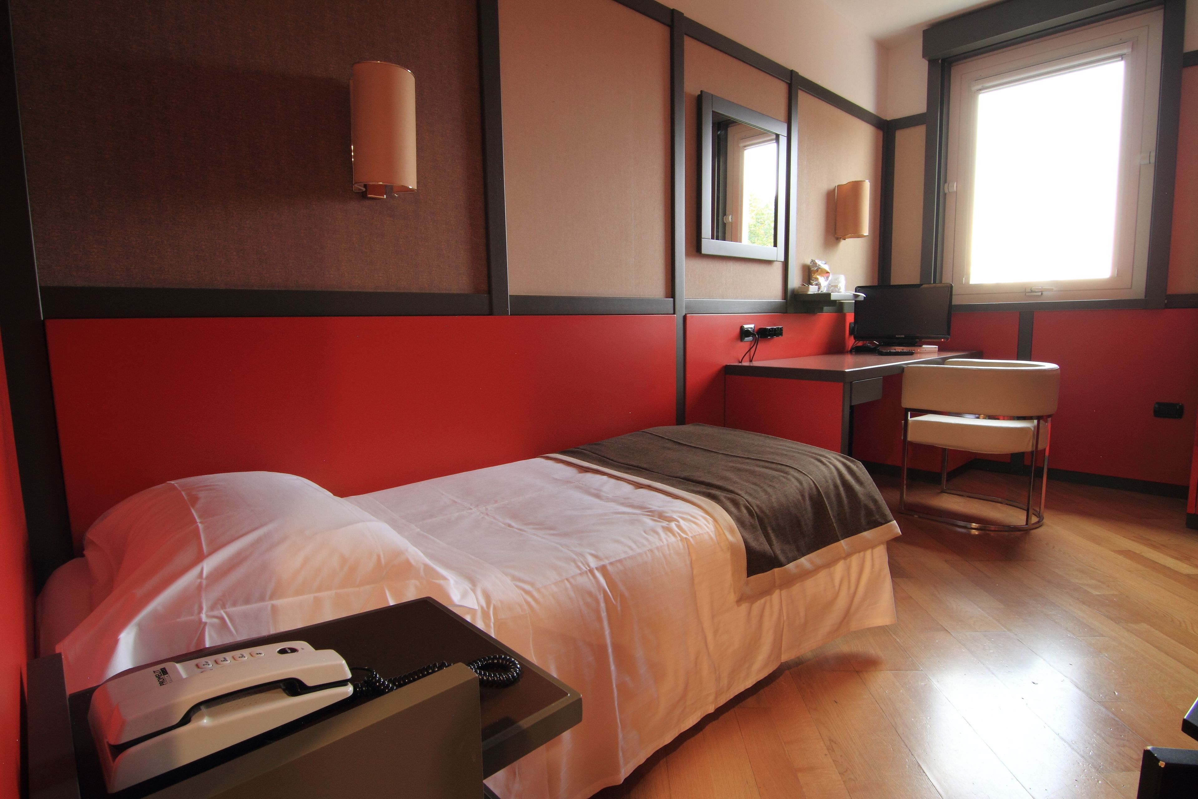 A hotel room with a single bed, a desk with a computer, a chair, and a window with natural light.