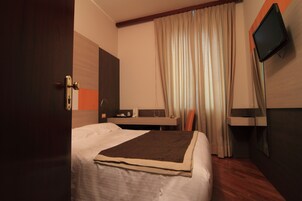 Una camera d'albergo con un letto, una scrivania, una sedia, una televisione e una finestra con tende.