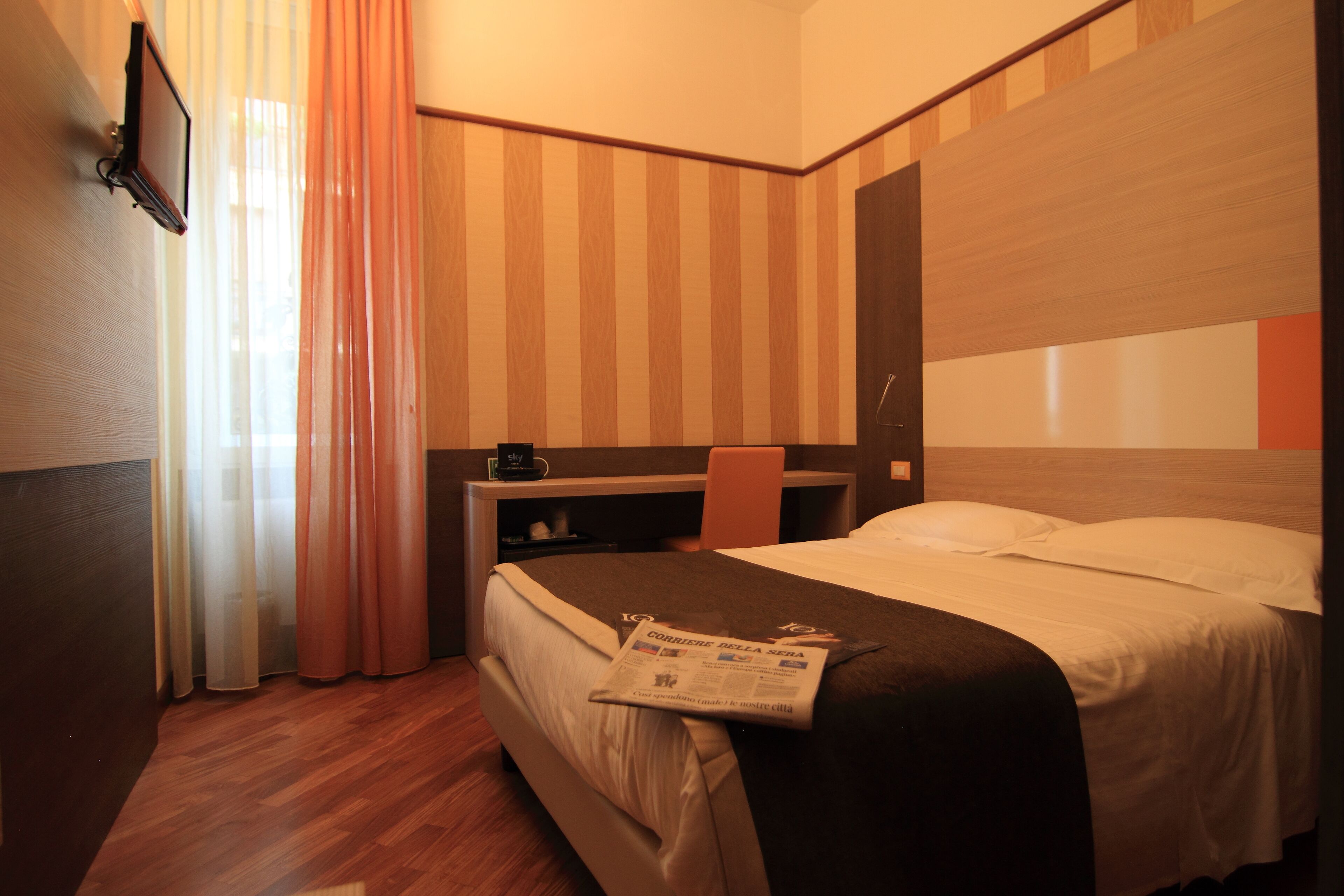 Ein Hotelzimmer mit Bett, Schreibtisch, Stuhl und einem an der Wand befestigten Fernseher.