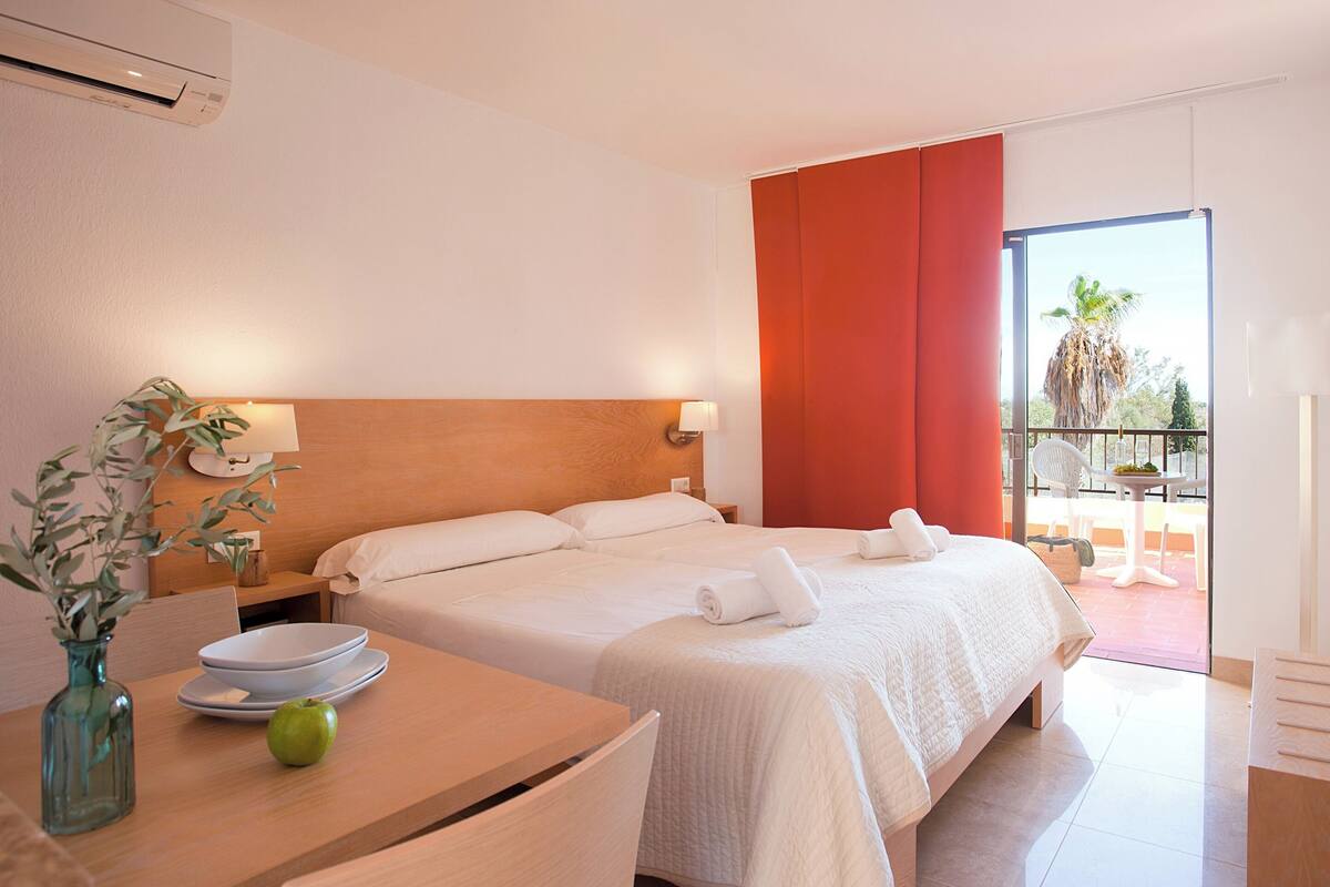 Une chambre d’hôtel avec un lit, une table ornée d’un vase et d’une pomme, un balcon offrant une vue sur les palmiers, et un rideau rouge.
