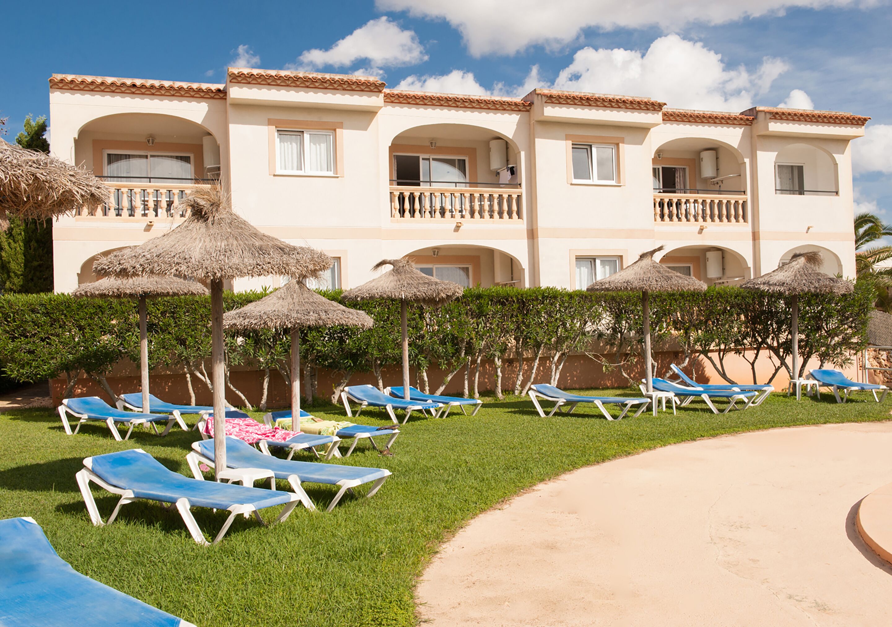 Un hotel con varios balcones, una zona de piscina con tumbonas y sombrillas, y un césped bien cuidado.