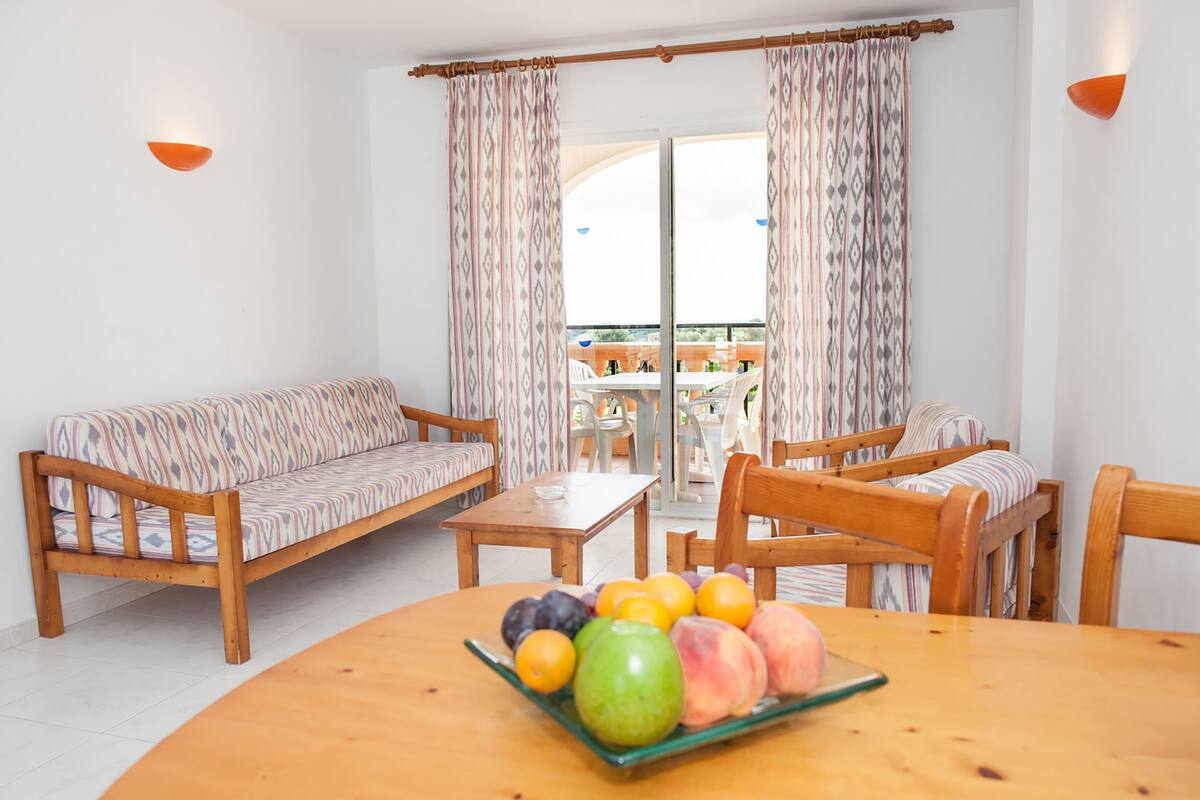 Une pièce avec un canapé, des fauteuils, une table avec des fruits et un balcon avec des rideaux.