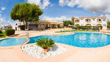 Una piscina con una zona hundida, rodeada de guijarros blancos y una planta de palmera. Hay sillas de descanso y sombrillas alrededor de la piscina.