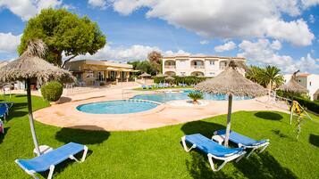 Un área de piscina con sillones de descanso, una estructura de techo de paja y un edificio con balcones al fondo.