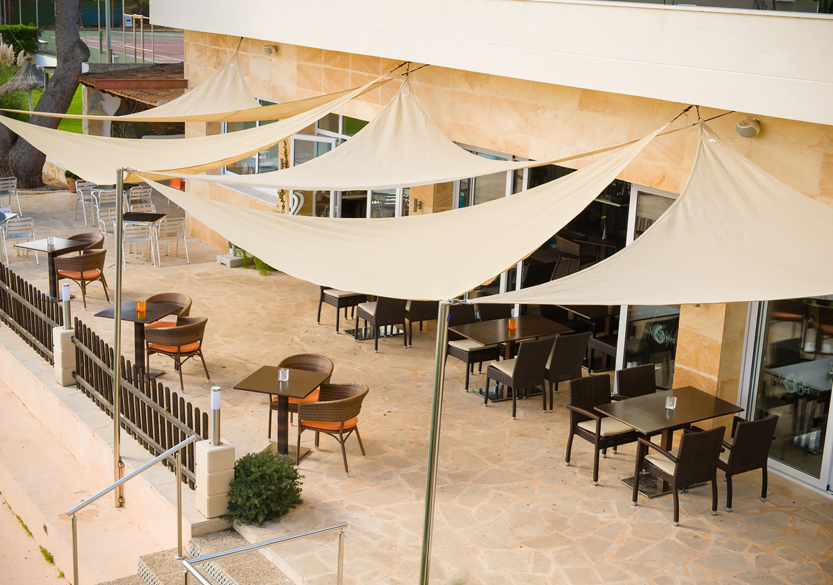 Een buitenpatio met tafels en stoelen, beschaduwd door grote zonnedaken, en een stenen vloer.