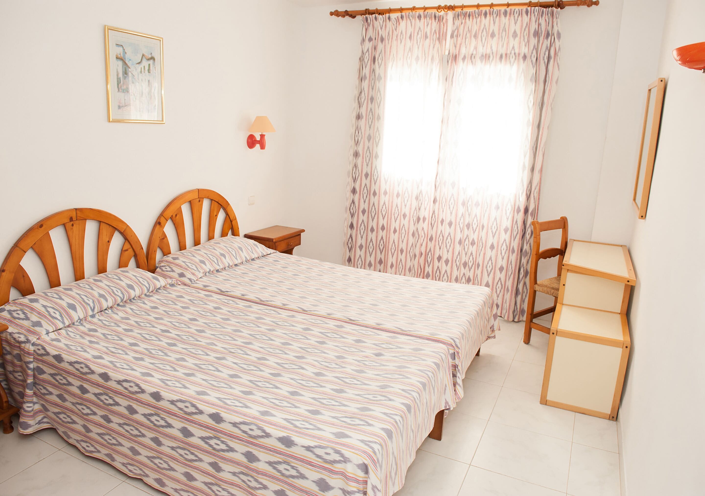Une chambre à coucher avec un lit, une chaise, une table de chevet, une lampe et une fenêtre avec des rideaux.