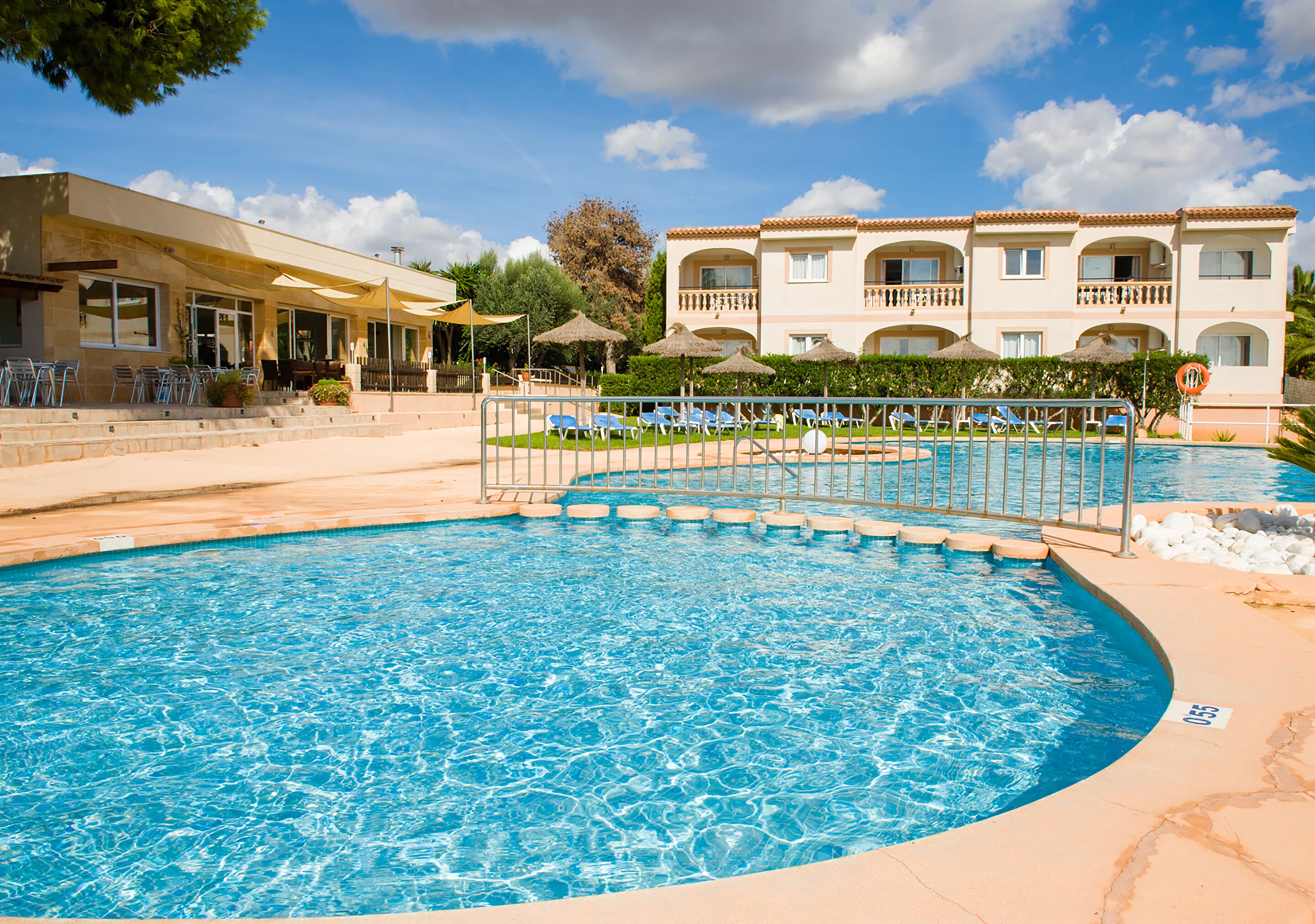 Una piscina con sillones de playa y sombrillas, un edificio con balcones y un cielo despejado.