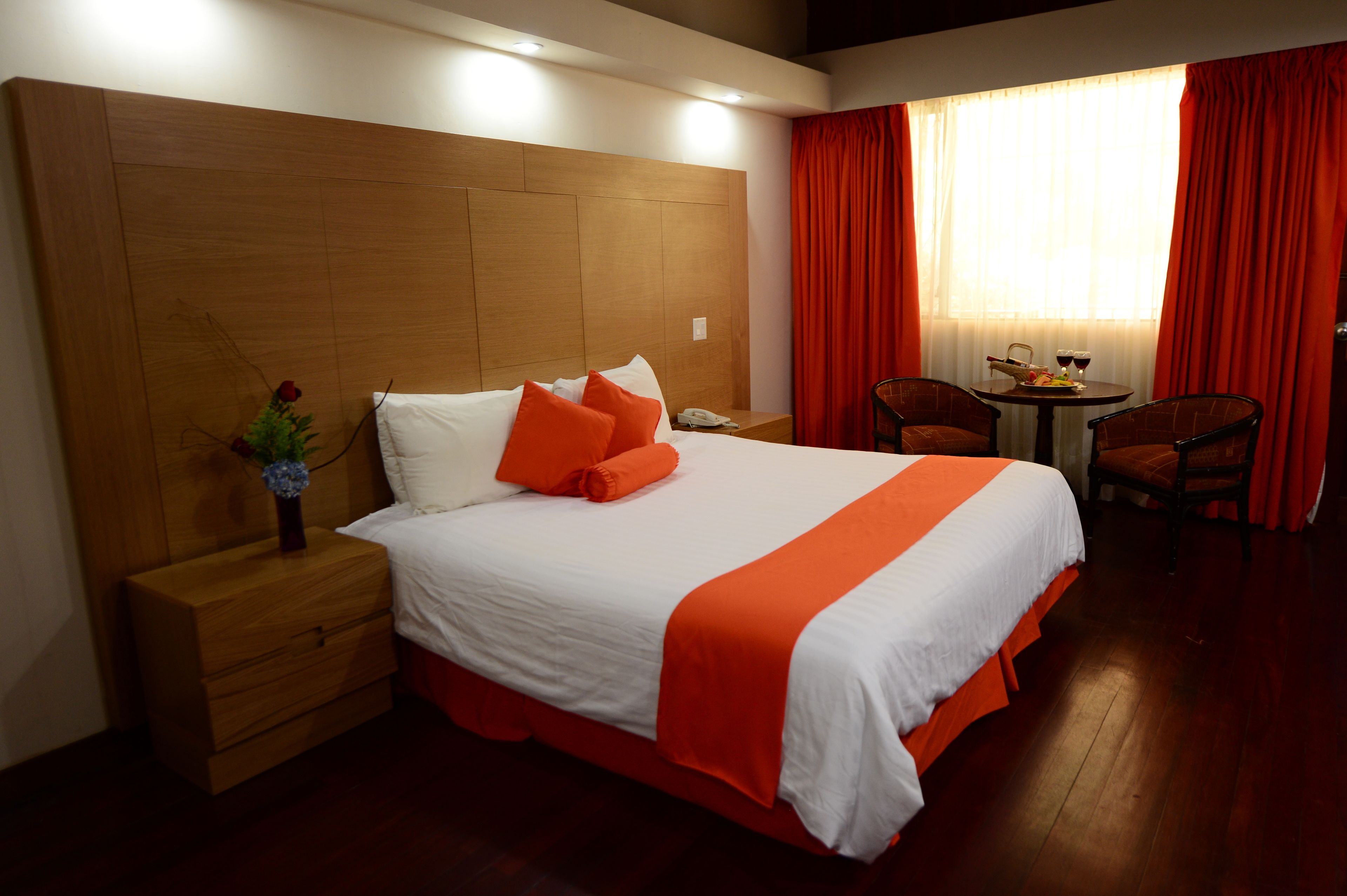 A hotel room with a large bed, wooden headboard, a nightstand, a small table with a chair, and a window with red curtains.