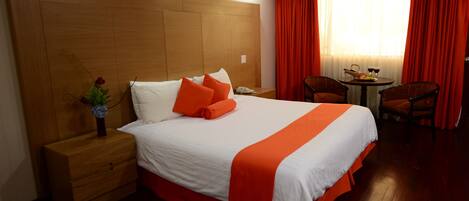 Habitación de hotel con una cama grande, cabecera de madera, una mesita de noche, una pequeña mesa con una silla y una ventana con cortinas rojas.