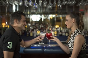 A bar setting with a man and a woman holding drinks, a bar counter, and a background of shelves with bottles.