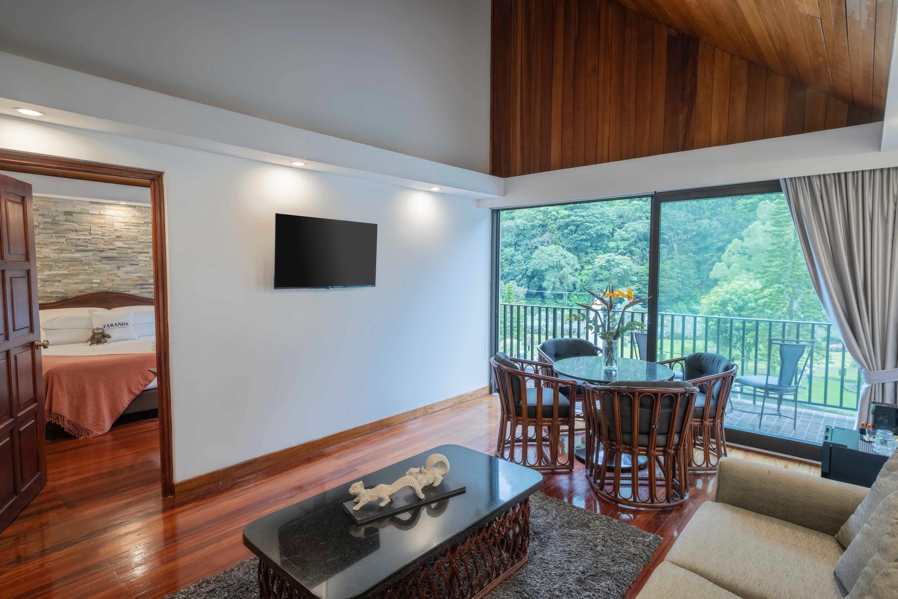 A spacious living area with a wooden floor, a flat-screen TV mounted on the wall, a dining table with chairs, and a sofa.