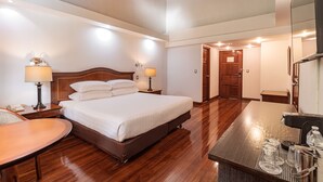 Habitación de hotel con una cama grande, lámparas de noche, suelo de madera, un escritorio con espejo y la puerta del baño.