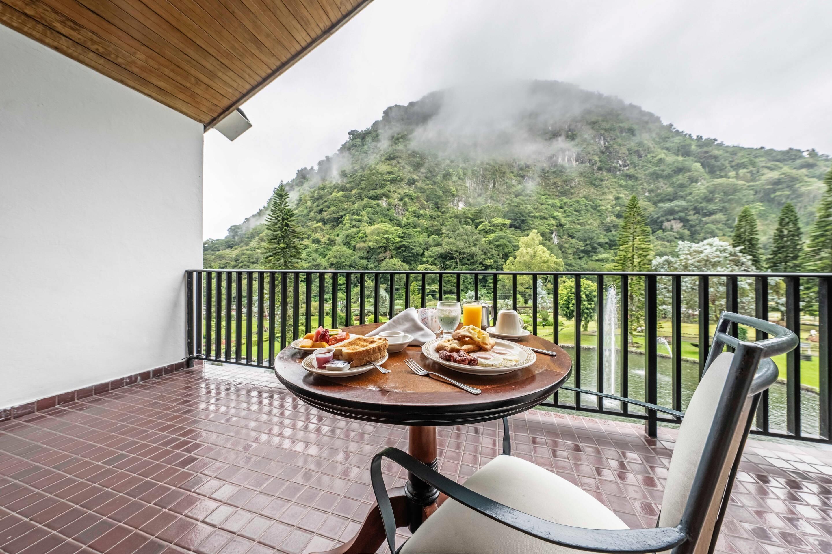 Un balcón con una mesa lista para el desayuno, con vistas a un paisaje verde y exuberante y una montaña envuelta en niebla.