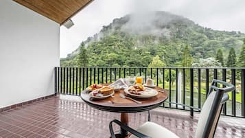 Un balcón con una mesa lista para el desayuno, con vistas a un paisaje verde y exuberante y una montaña envuelta en niebla.