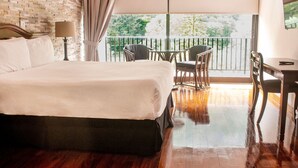 Habitación de hotel con una cama grande, un escritorio y sillas, un balcón con vistas a zonas verdes y una pared de ladrillo.