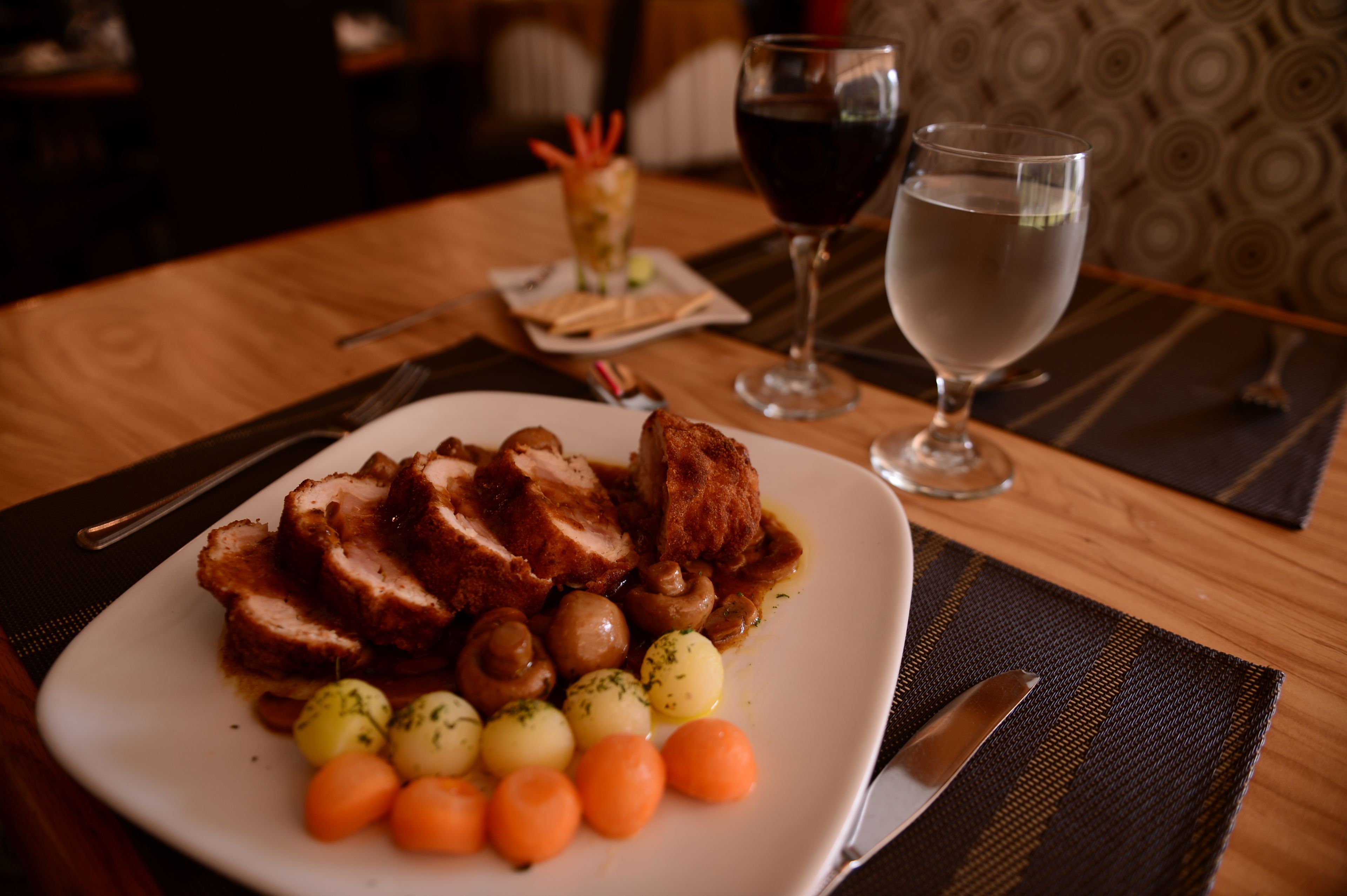 Ein Teller mit geschnittenem Fleisch, Pilzen und kleinen Gemüsesorten auf einer weißen Platte, dazu ein Glas Rotwein und ein Glas klares Wasser auf einem Holztisch.