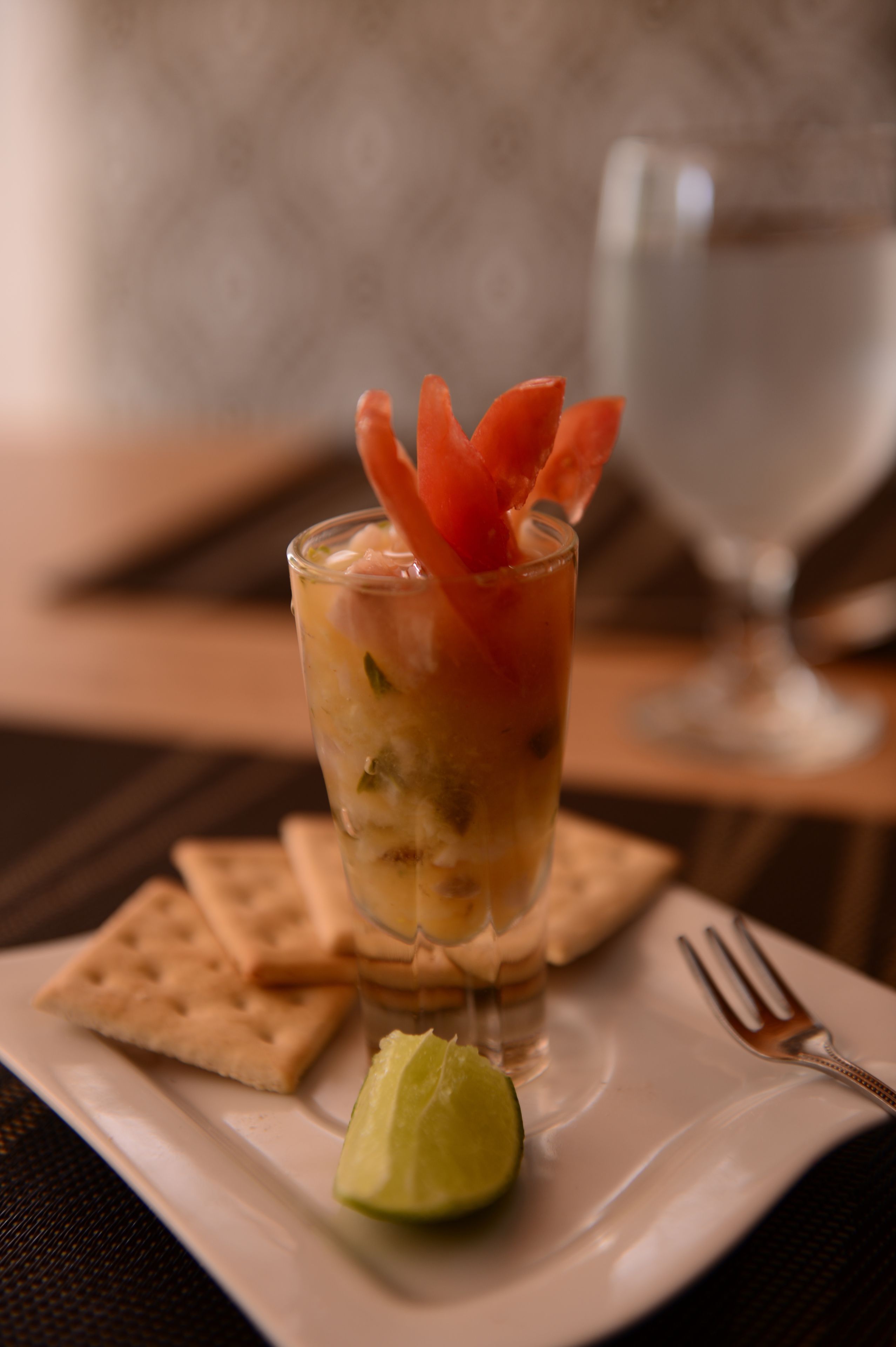 A glass of cocktail with a lime wedge and carrot garnish on a white plate, accompanied by crackers and a fork.