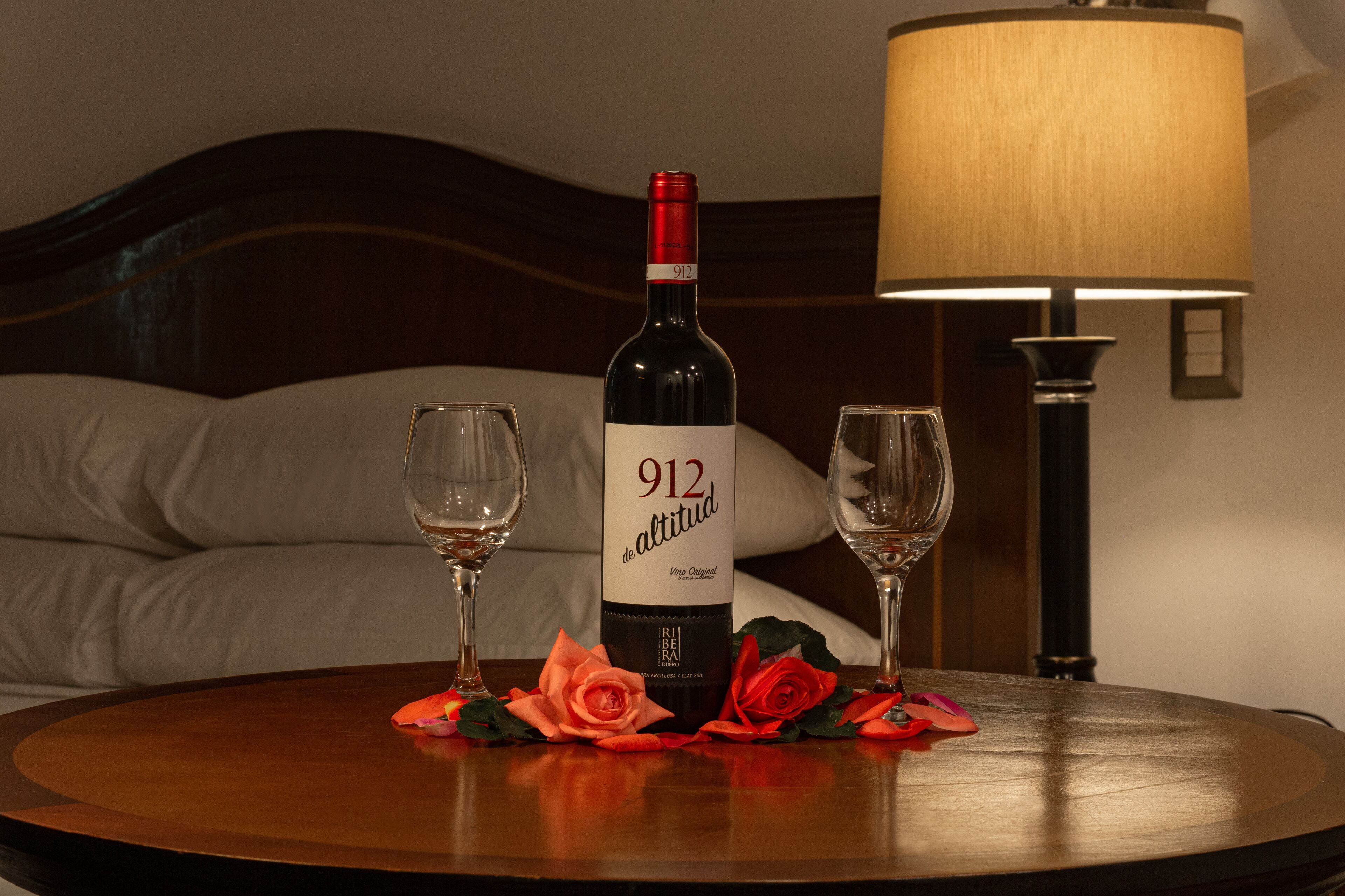 A bottle of 912 de Altitud wine, two glasses, and decorative roses on a round table in a hotel room.