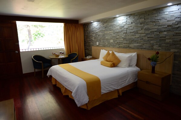 A hotel room with a large bed, a wooden nightstand, a small table with a chair, and a window with curtains.