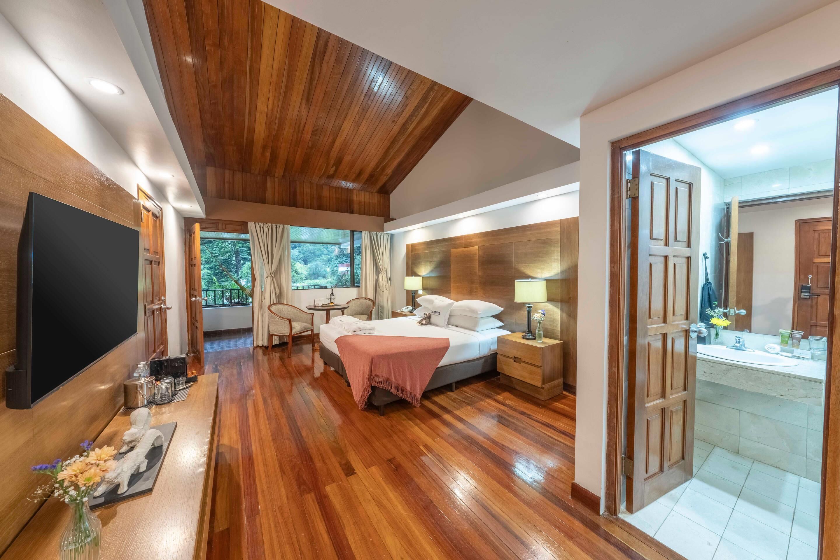 A hotel room with a wooden ceiling, a bed with a pink blanket, a TV, a desk with a lamp, and a bathroom with a mirror and sink.