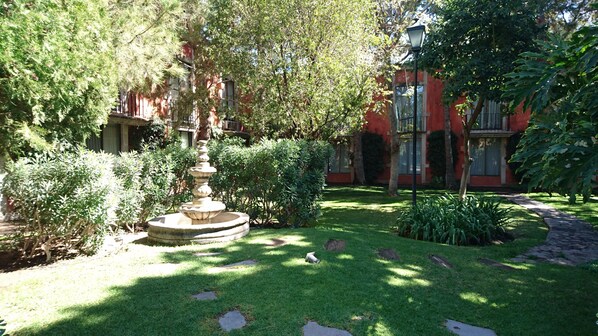 A garden with a stone fountain, lush greenery, and a red building in the background.