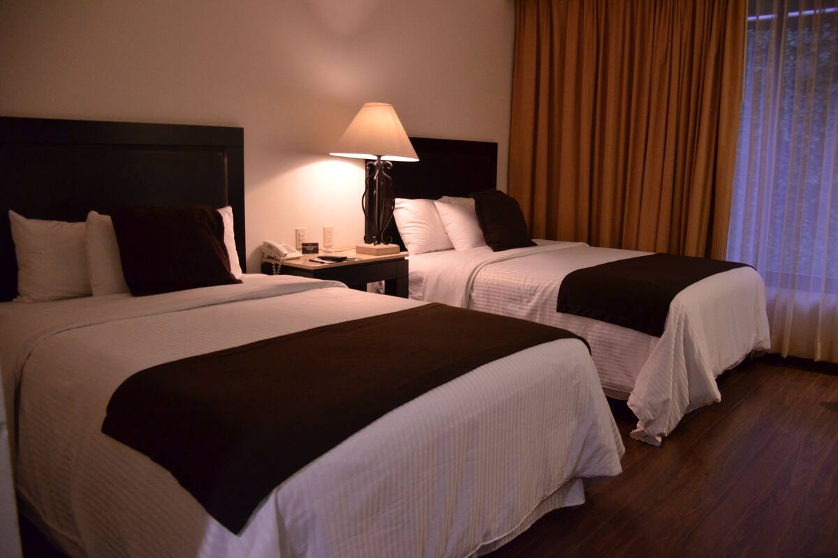 Una habitación de hotel con dos camas, una mesita de noche con lámpara y una ventana con cortinas.