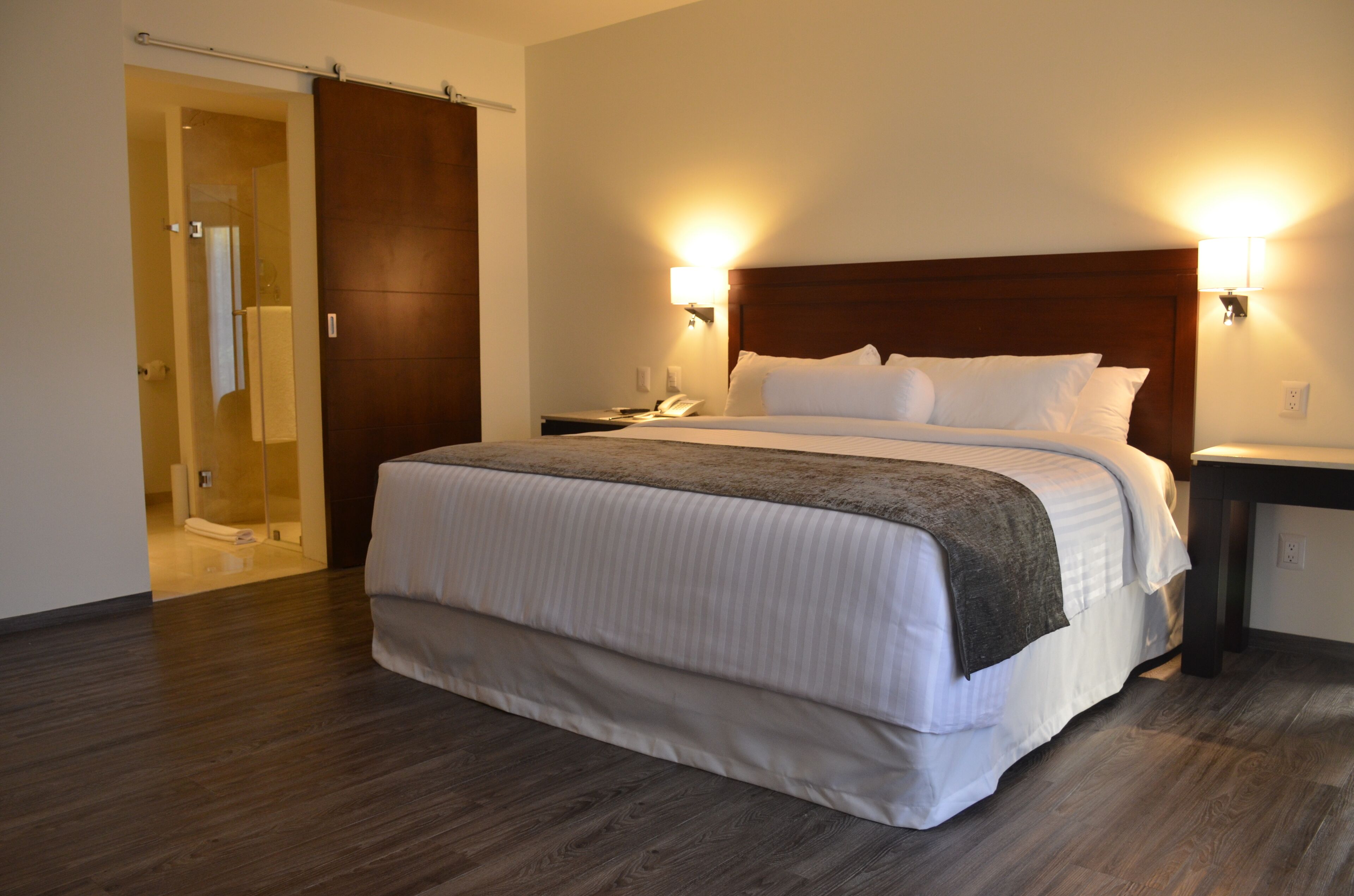 A hotel room with a large bed, wooden headboard, two bedside tables, a nightstand, and a sliding door leading to a bathroom.