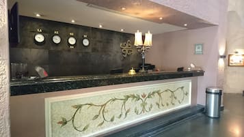 Una recepción con relojes que muestran diferentes zonas horarias, un mosaico decorativo y un cubo de basura.