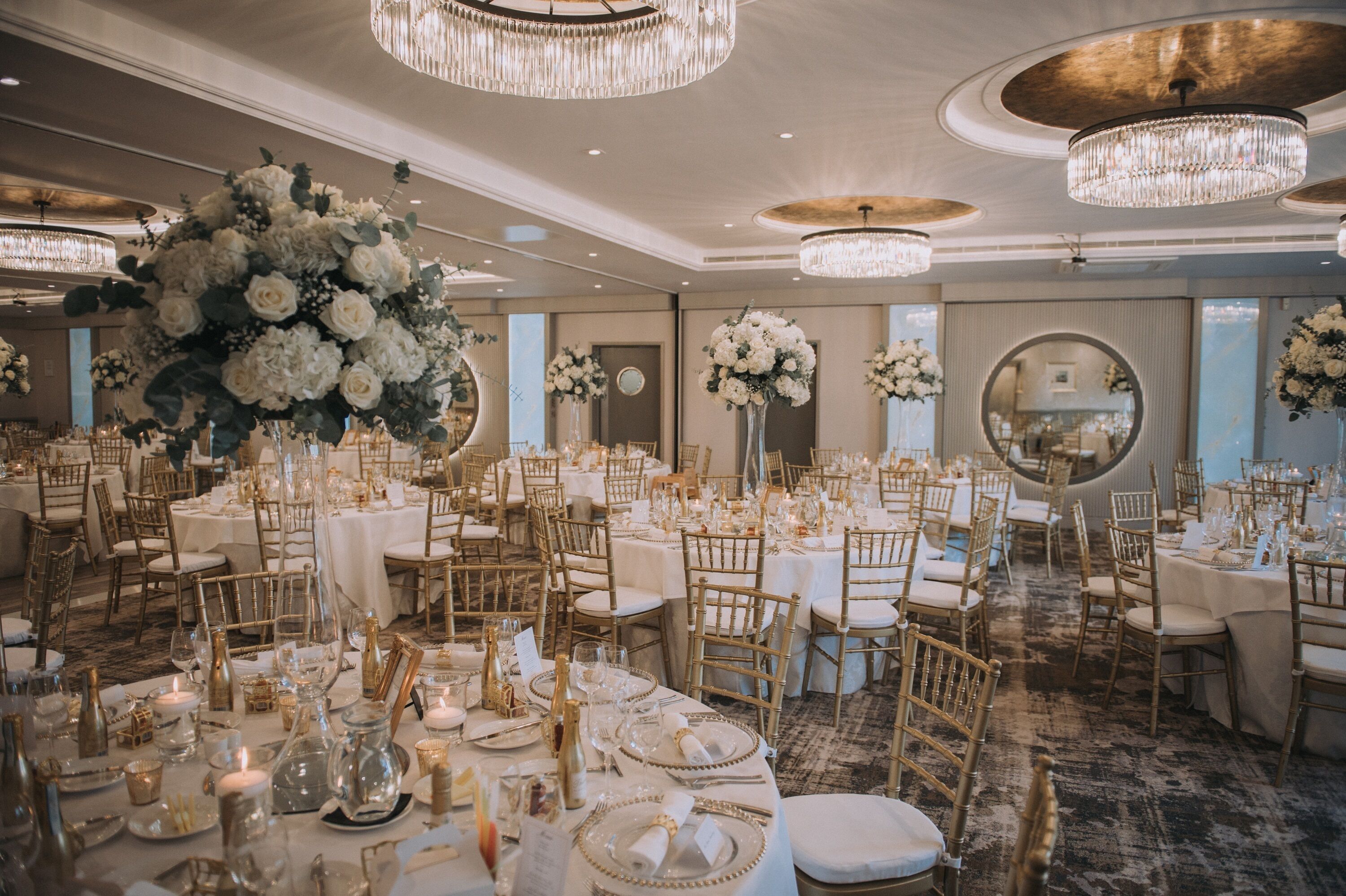 A banquet hall with round tables, gold chairs, and large floral arrangements.