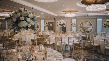 A banquet hall with round tables, gold chairs, and large floral arrangements.