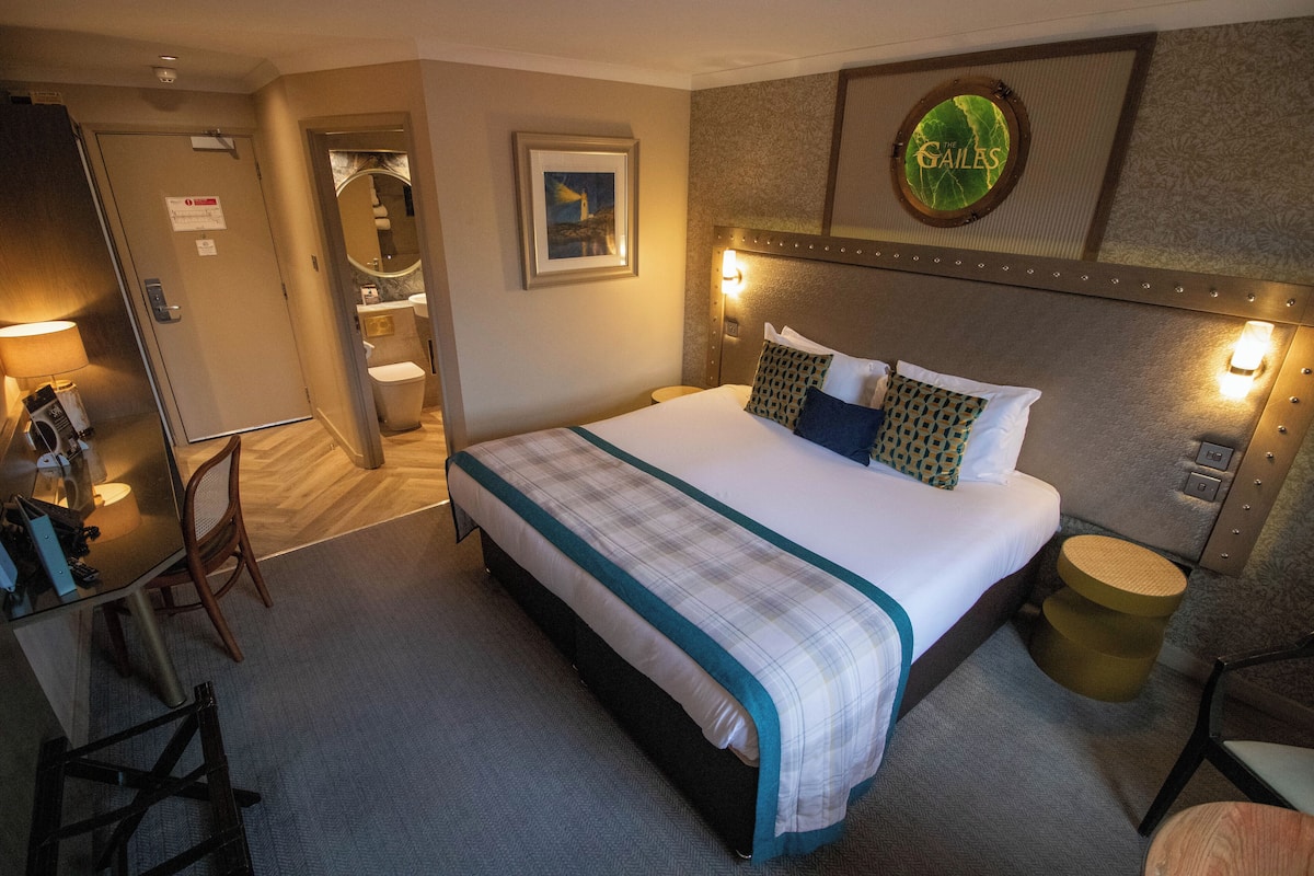 A hotel room with a large bed, a desk, a chair, a round side table, and a bathroom visible through an open door.