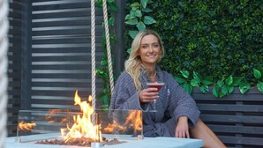 A person sitting by a glass table with a fire, in front of a wooden fence and a green leafy wall. - The Gailes Hotel and Spa (Irvine)