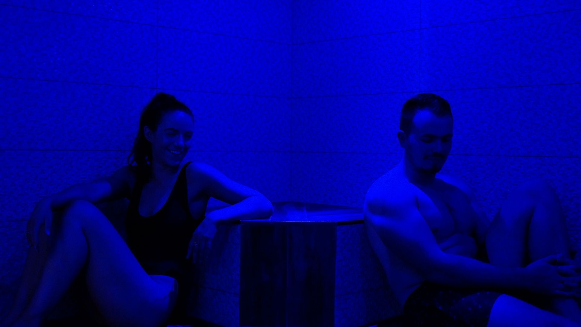 Two people sitting on a bench in a room with a tiled wall.