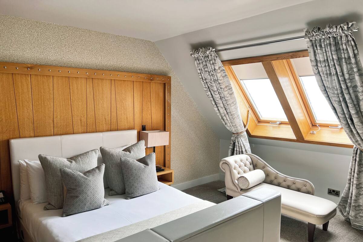 A bedroom with a bed, bedside table, lamp, chair, and a window with curtains.