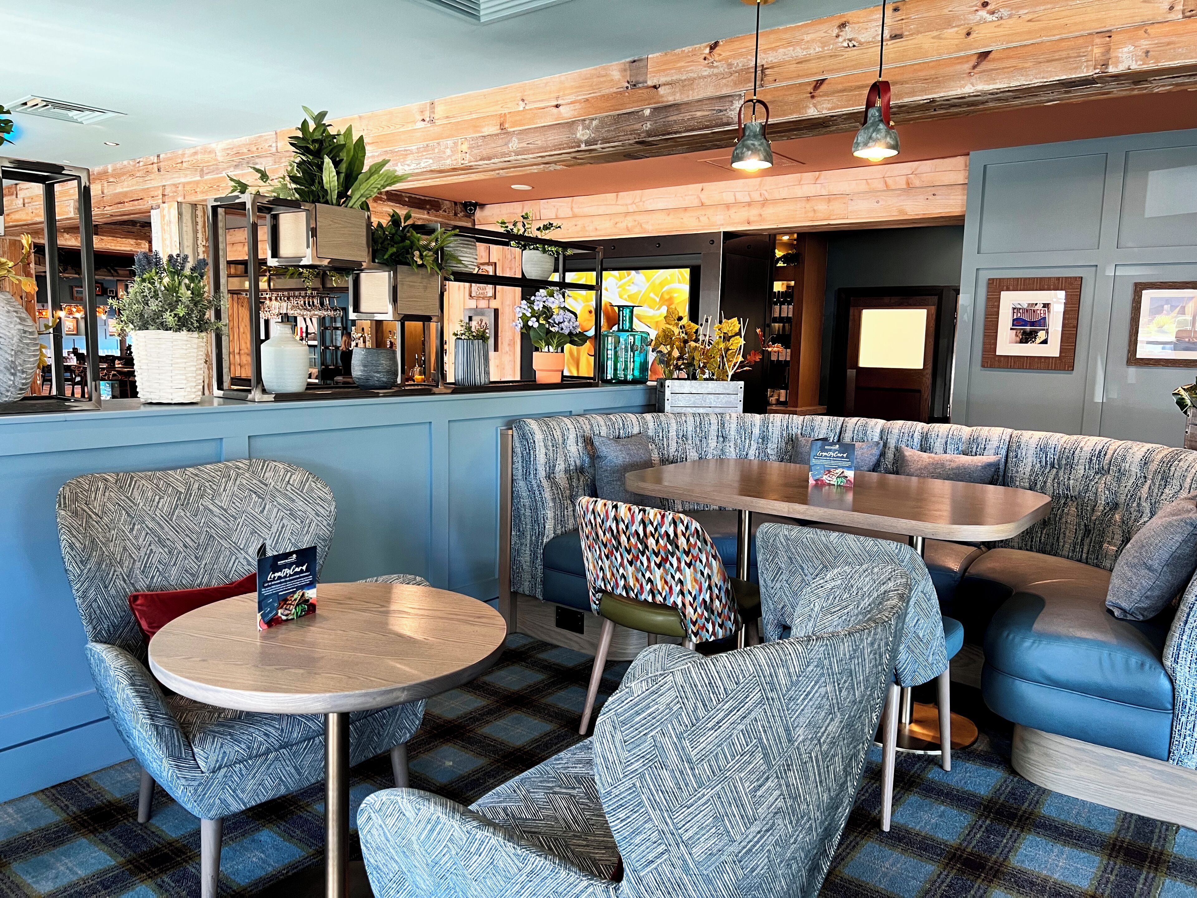 A cozy seating area with patterned chairs and a round table, a bar area with plants and bottles, and a wooden ceiling.