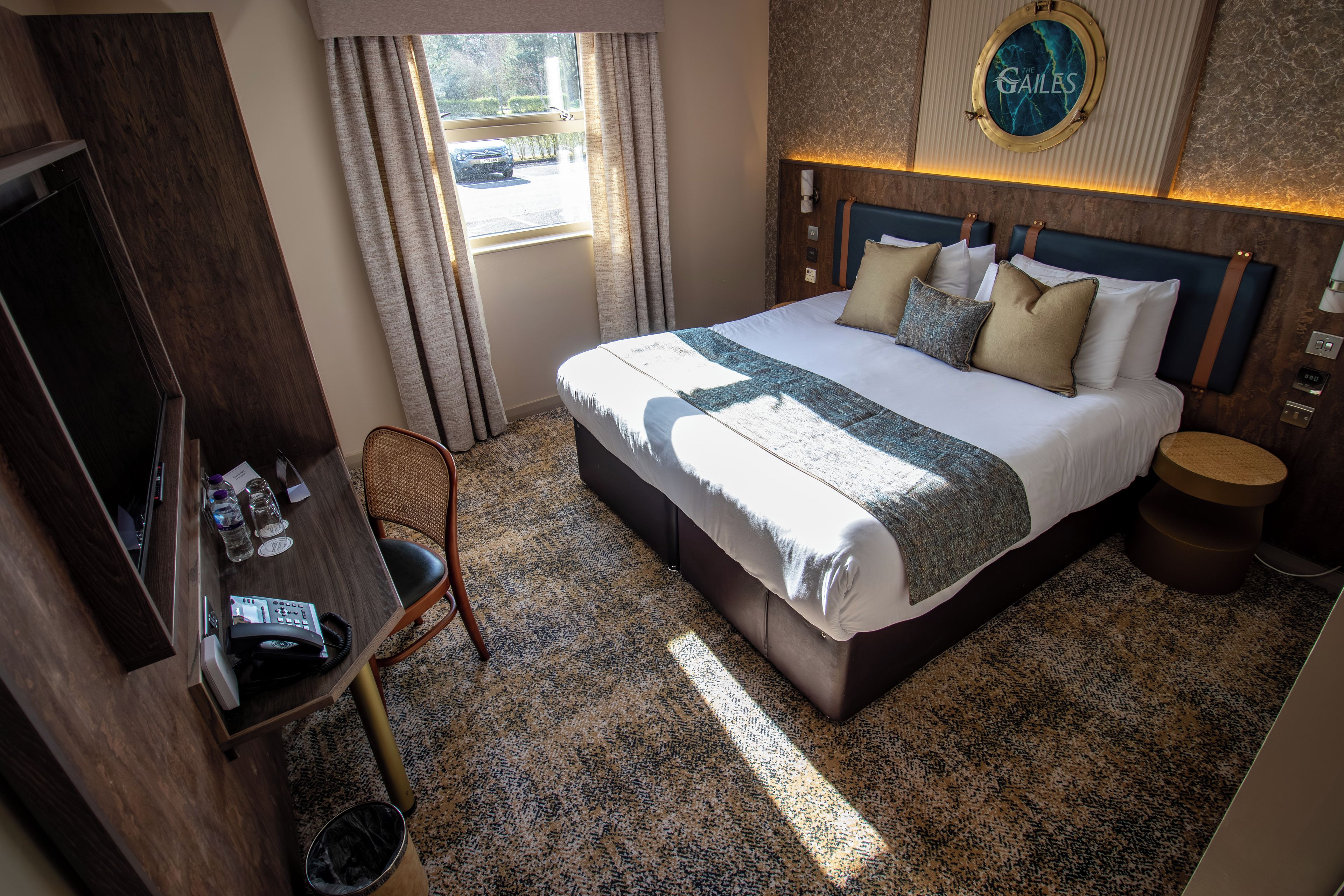 Una habitación de hotel con cama, escritorio, silla, cubo de basura y ventana con cortinas.