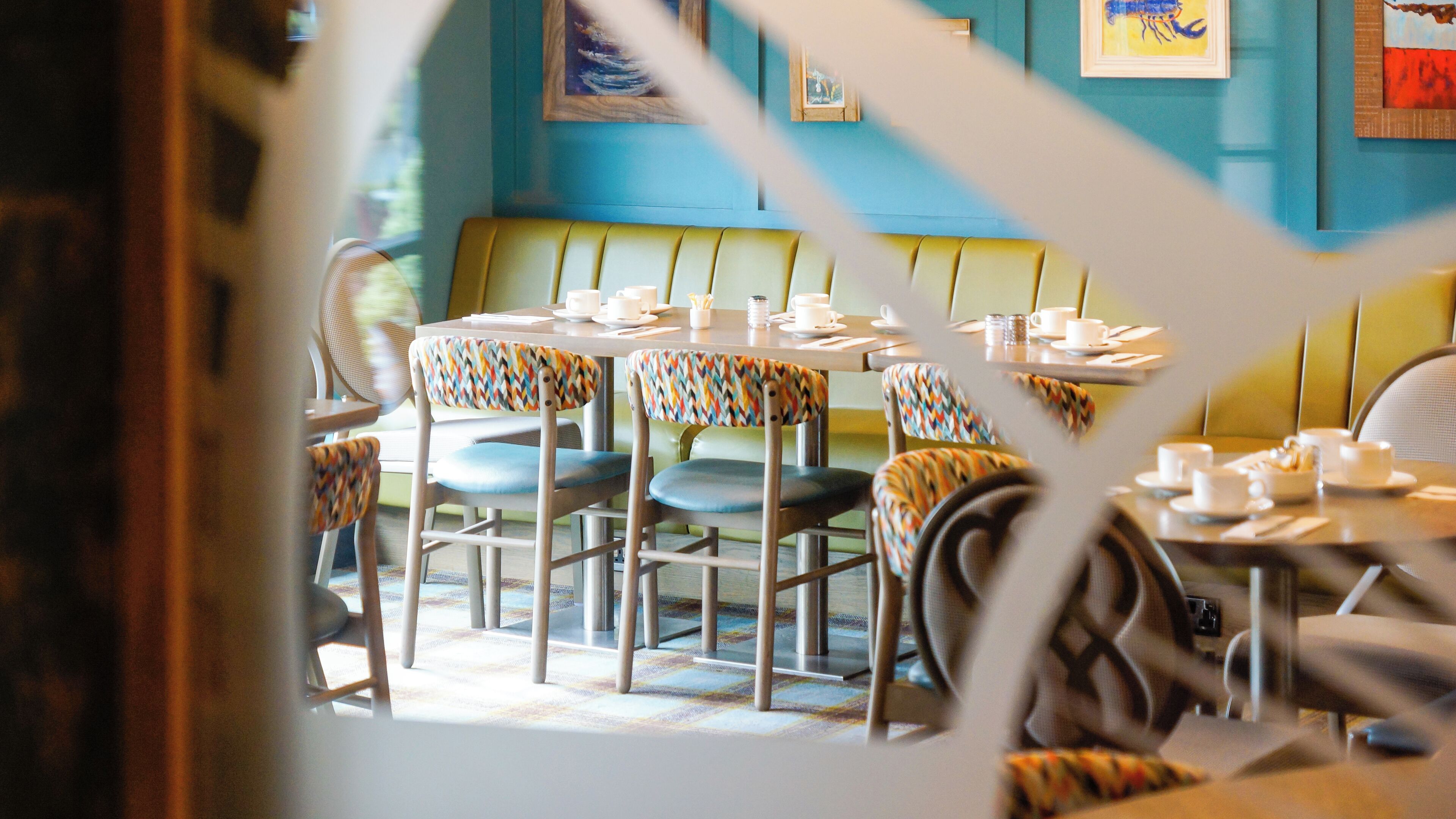 Un interior de restaurante con mesas redondas, sillas de colores con diseños y una pared azul decorada con obras de arte.
