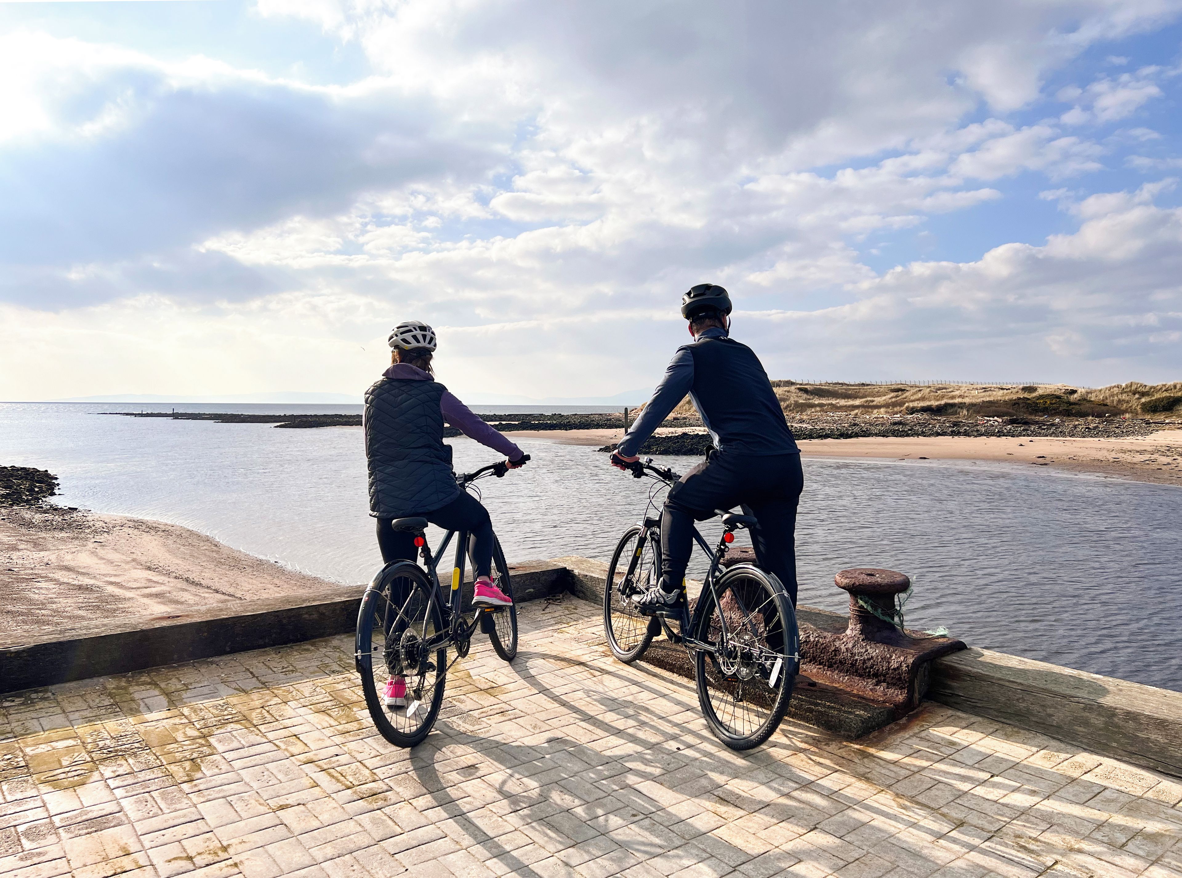 Two cyclists on a paved path by a body of water with a distant shoreline.