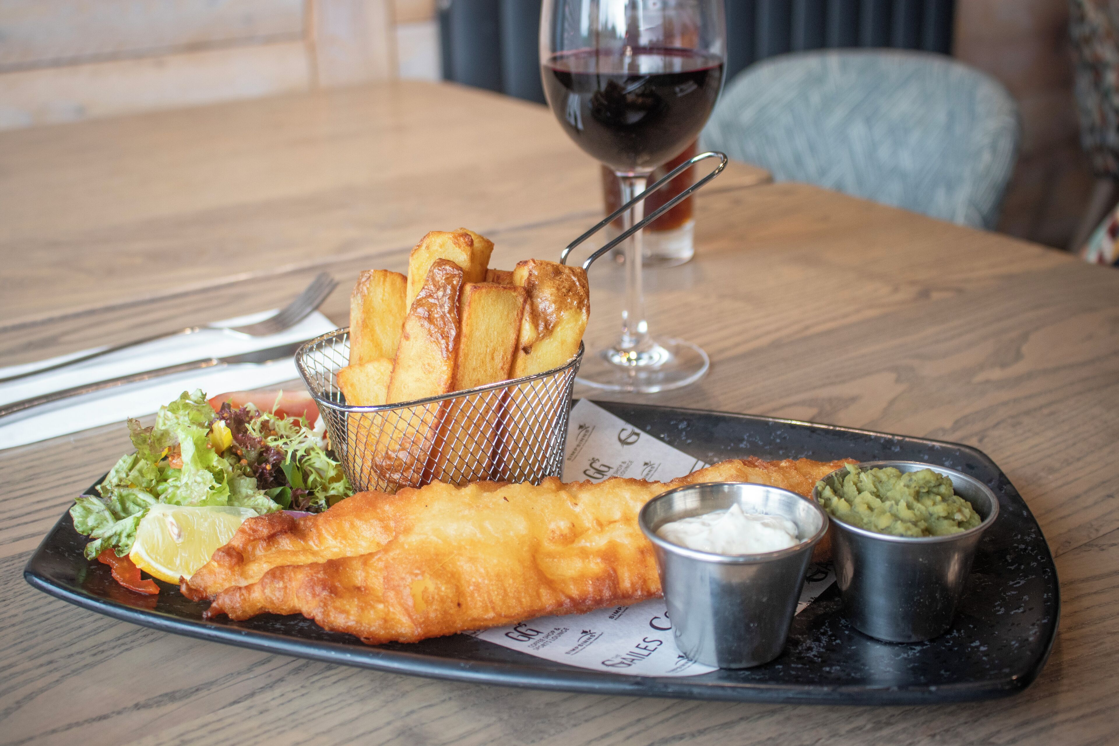 A plate of fish and chips with a side salad, chips, and two dipping sauces on a wooden table.