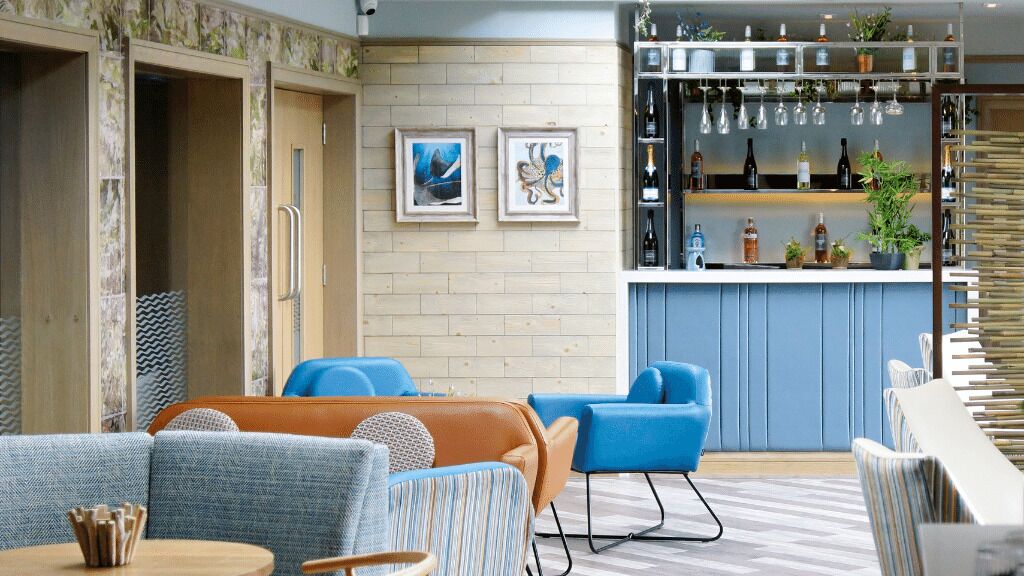Un espace lounge moderne avec des sièges bleus et bruns, un bar doté d’étagères remplies de bouteilles et des œuvres d’art encadrées accrochées au mur.