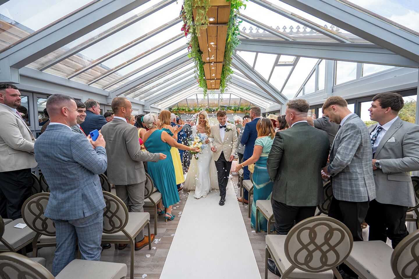 A wedding ceremony in a covered outdoor venue with a glass roof, seating arrangements, and hanging plants.