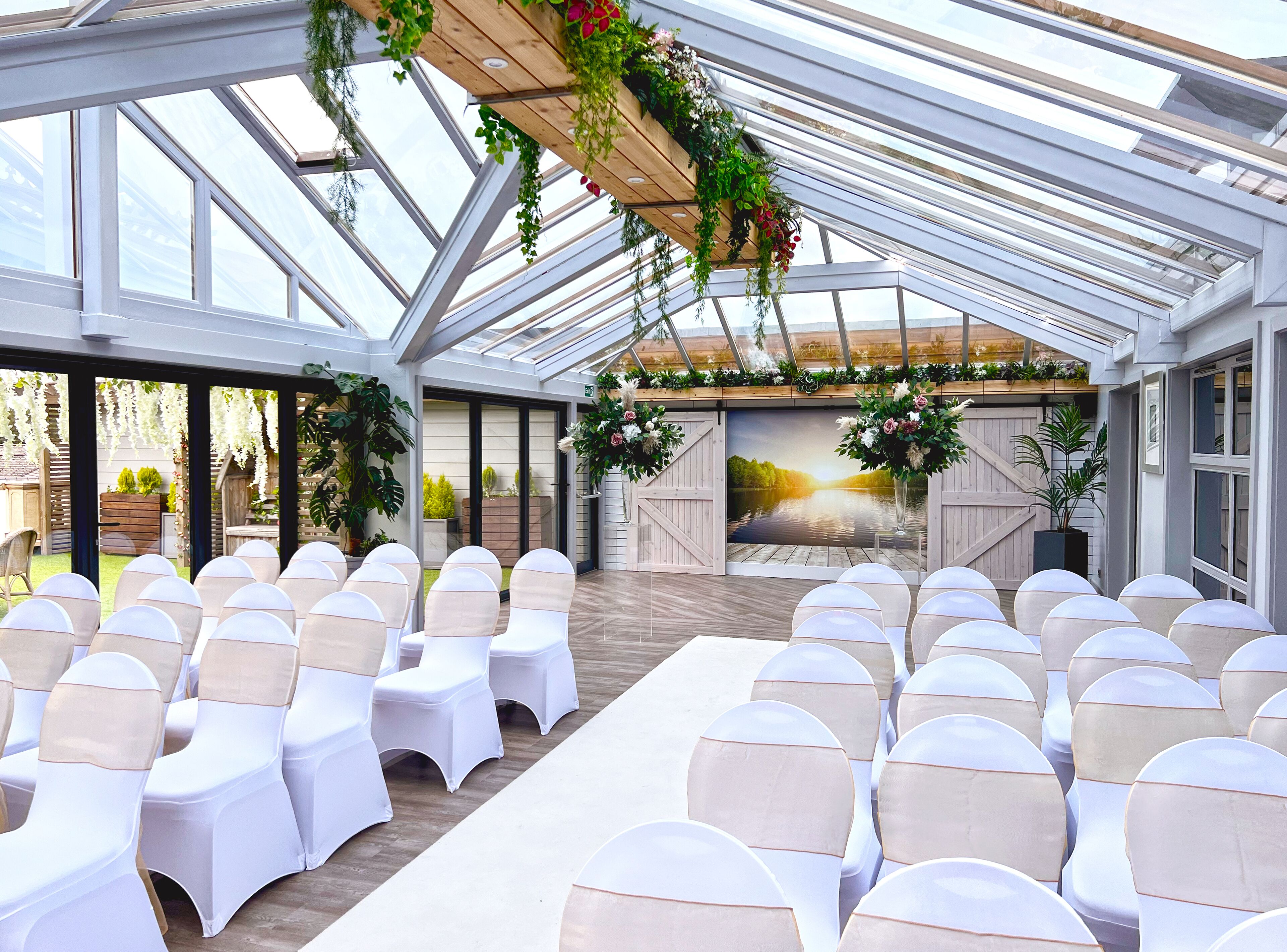 A spacious indoor venue with a high ceiling and large windows, featuring rows of white chairs with beige sashes, a wooden stage with floral decorations, and a scenic mural on the back wall.