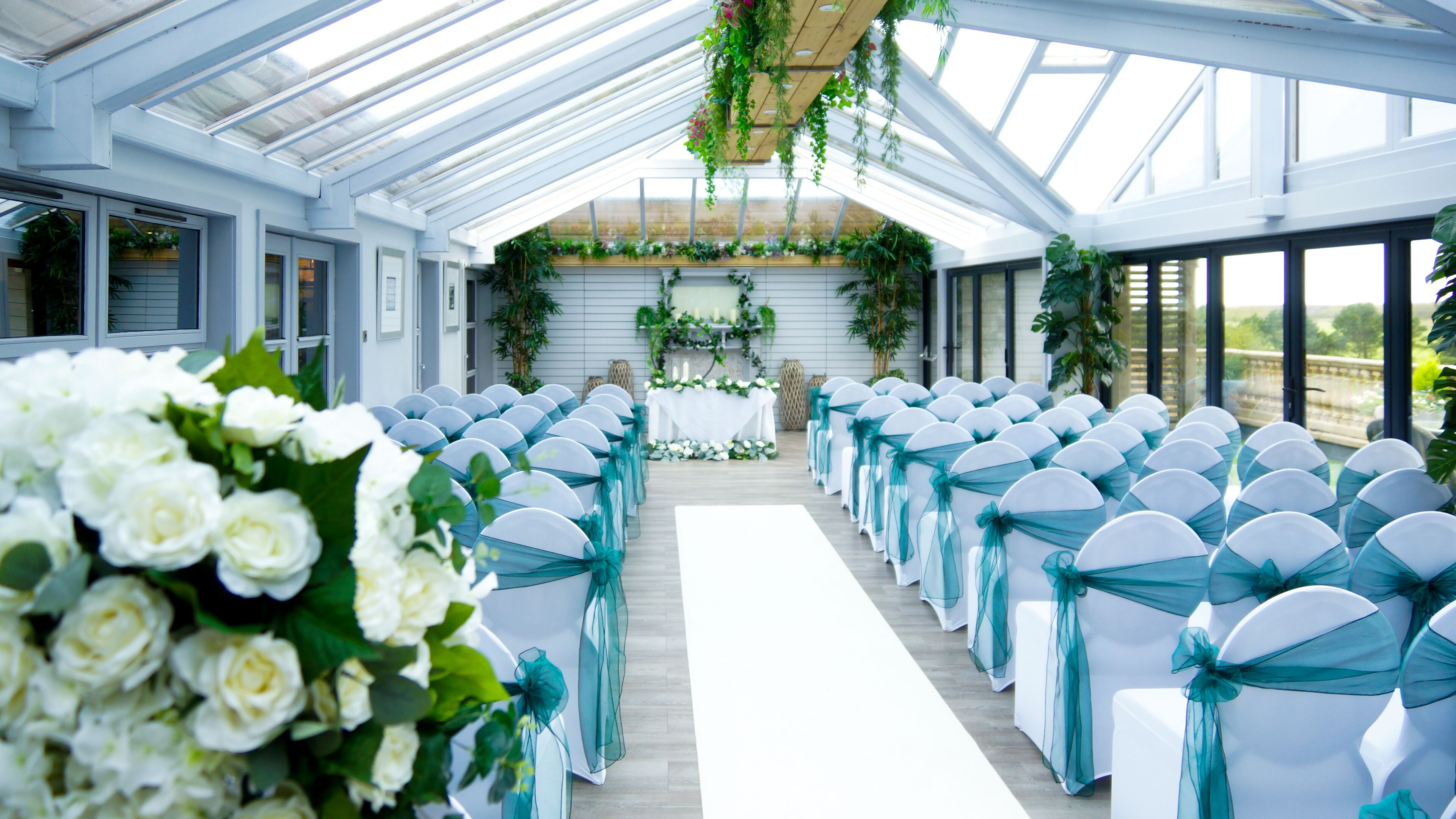 A covered aisle with white chairs and teal sashes, a floral arrangement, and a table with greenery and candles.
