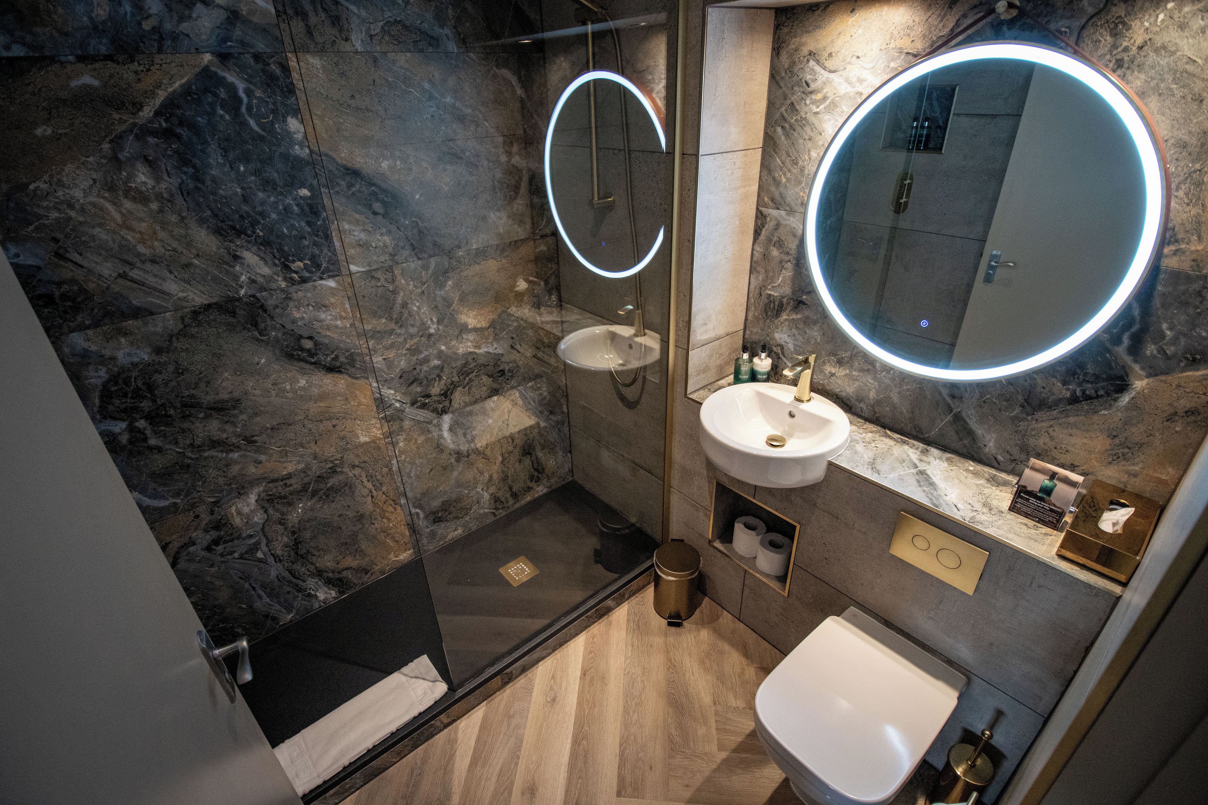 Un baño moderno con pared de mármol, ducha con cerramiento de vidrio, lavamanos blanco y espejo redondo con luz.
