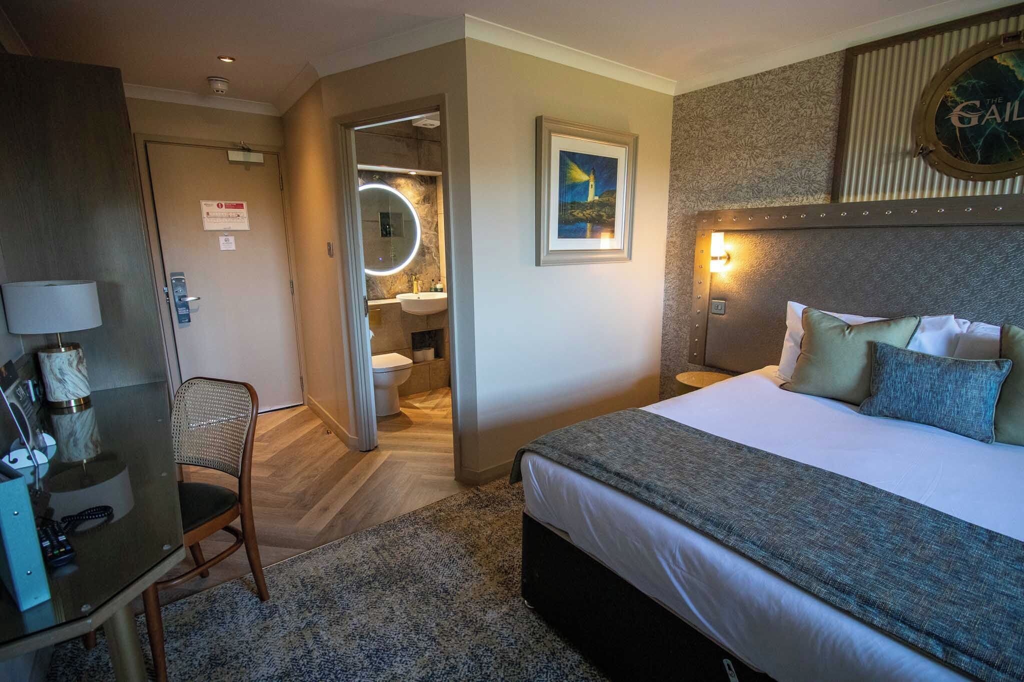 A hotel room with a bed, a desk, a chair, a bathroom, and a wall-mounted clock.