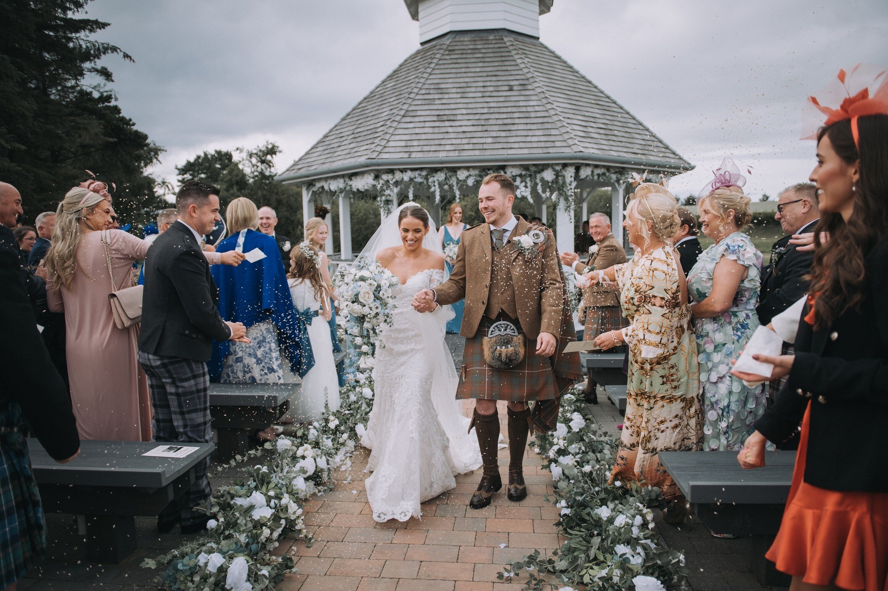 A wedding ceremony with guests seated on benches, a gazebo in the background, and a floral aisle.