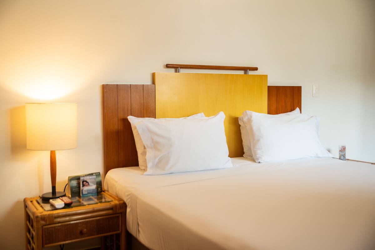 A hotel room with a bed, a bedside table with a lamp, and a nightstand with a remote control and a magazine.