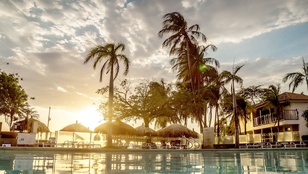 Zona de piscina con sillones de descanso y sombrillas, rodeada de palmeras y un edificio con techo de tejas.