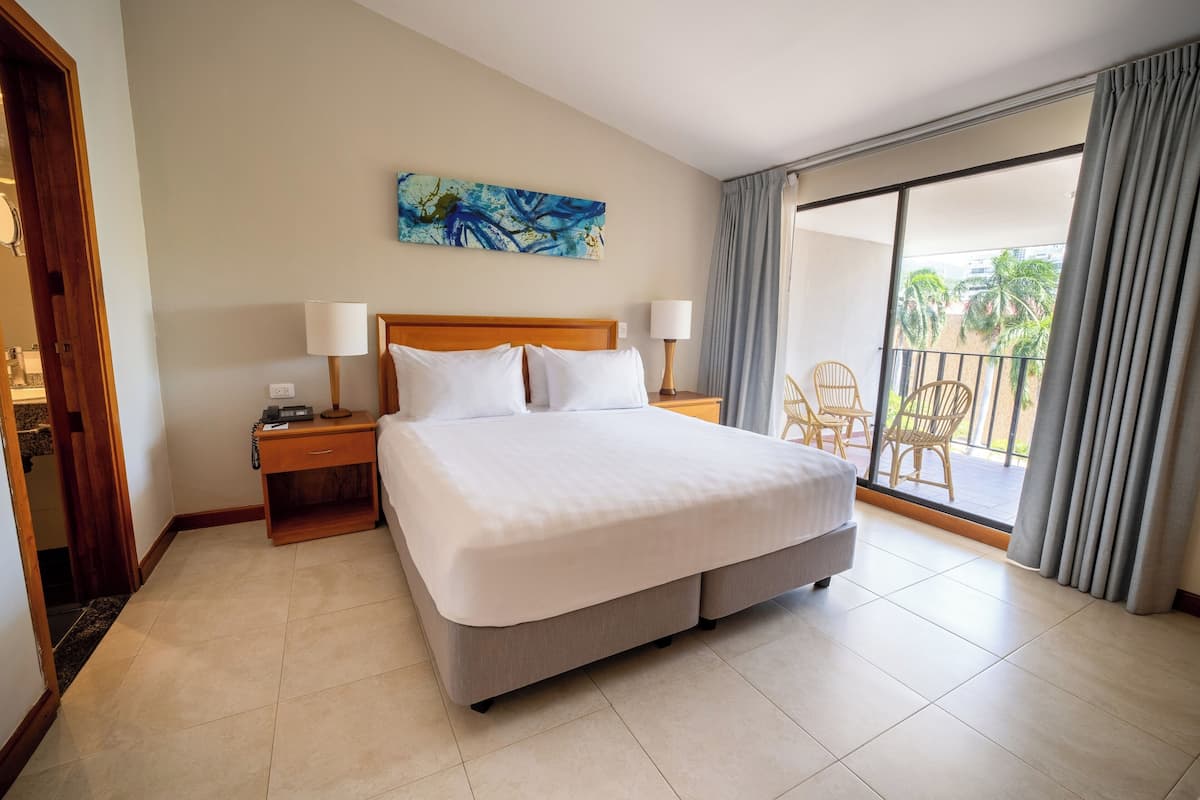 A hotel room with a large bed, two bedside lamps, a nightstand, a painting on the wall, and a balcony with chairs.
