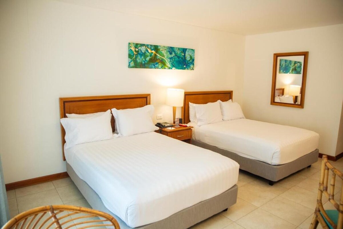 A hotel room with two beds, a nightstand with a lamp, a chair, and a painting on the wall.